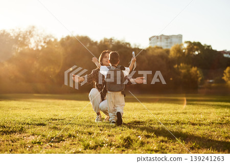 Pupil is with book and backpack. Mother and son together in an open field during daytime Pupil is with book and backpack. Mother and son together in an open field during daytime 139241263
