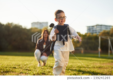 Walking forward, pupil is with book and backpack. Mother and son together in an open field during daytime 139241265