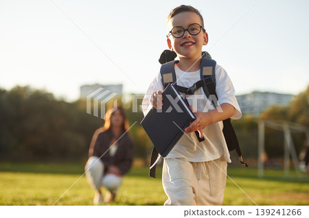 Walking forward, pupil is with book and backpack. Mother and son together in an open field during daytime Walking forward, pupil is with book and backpack. Mother and son together in an open field during daytime 139241266