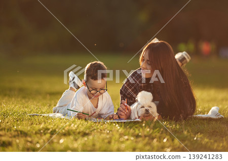 Relaxed, on the blanket, with little cute dog. Mother and son together in an open field during daytime Relaxed, on the blanket, with little cute dog. Mother and son together in an open field during daytime 139241283
