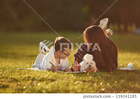 Dog is lying down with owners that are drawing. Mother and son together in an open field during daytime 139241285