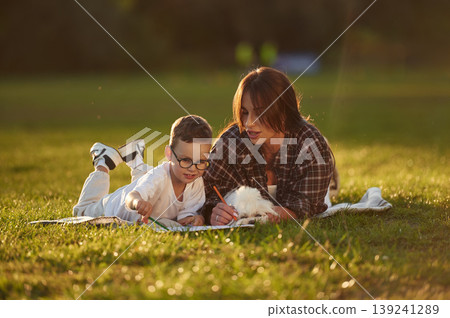 Dog is lying down with owners that are drawing. Mother and son together in an open field during daytime Dog is lying down with owners that are drawing. Mother and son together in an open field during daytime 139241289