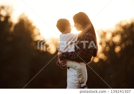 Woman is holding kid in hands, illuminated by sunlight. Mother and son together in an open field during daytime Woman is holding kid in hands, illuminated by sunlight. Mother and son together in an open field during daytime 139241307