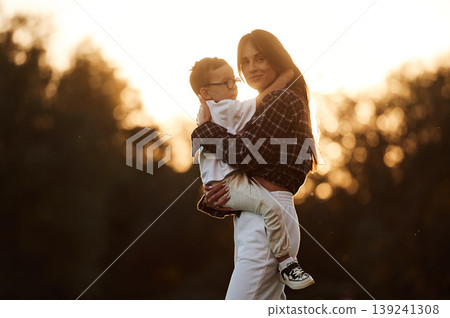 Woman is holding kid in hands, illuminated by sunlight. Mother and son together in an open field during daytime Woman is holding kid in hands, illuminated by sunlight. Mother and son together in an open field during daytime 139241308