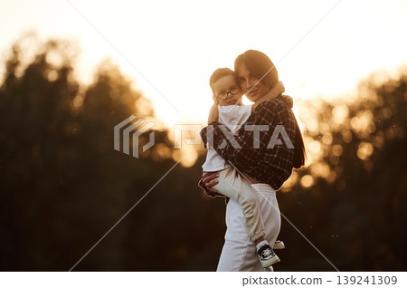 Woman is holding kid in hands, illuminated by sunlight. Mother and son together in an open field during daytime 139241309