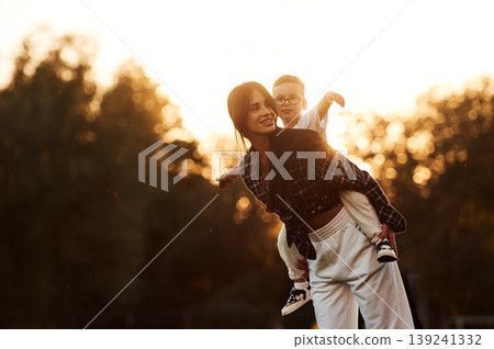 Trees illuminated by evening sunlight, background. Mother and son together in an open field during daytime Trees illuminated by evening sunlight, background. Mother and son together in an open field during daytime 139241332