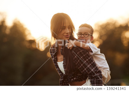 Positive facial expression, holding kid on back, sunset. Mother and son together in an open field during daytime 139241341