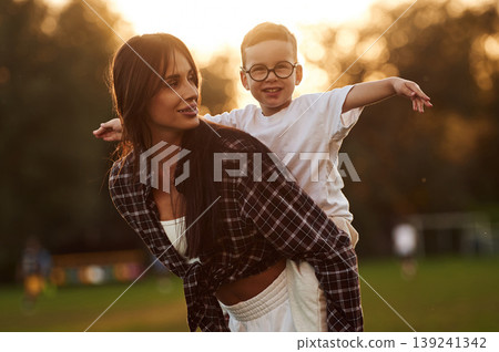 Positive facial expression, holding kid on back, sunset. Mother and son together in an open field during daytime Positive facial expression, holding kid on back, sunset. Mother and son together in an open field during daytime 139241342