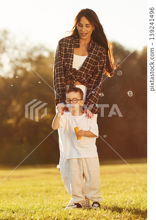 Front view, blowing bubbles. Mother and son together in an open field during daytime Front view, blowing bubbles. Mother and son together in an open field during daytime 139241496