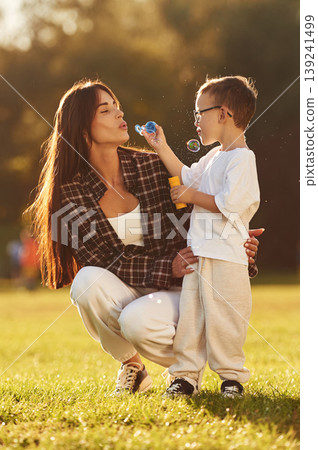 Front view, blowing bubbles. Mother and son together in an open field during daytime Front view, blowing bubbles. Mother and son together in an open field during daytime 139241499