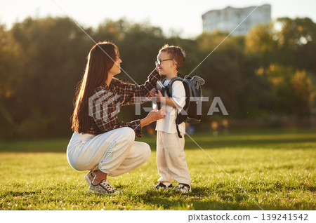 Getting ready for school. With book and backpack. Mother and son together in an open field during daytime Getting ready for school. With book and backpack. Mother and son together in an open field during daytime 139241542