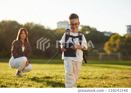 Pupil is with book and backpack. Mother and son together in an open field during daytime Pupil is with book and backpack. Mother and son together in an open field during daytime 139241548