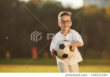 Holding soccer ball, front view. Boy is having fun on the field under sunlight at daytime Holding soccer ball, front view. Boy is having fun on the field under sunlight at daytime 139241584