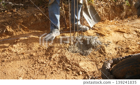 worker pours concrete into hole, rural construction site with concrete and rebar installation activities, scene depicting worker pouring wet concrete around rebar at rural construction site worker pours concrete into hole, rural construction site with concrete and rebar installation activities, scene depicting worker pouring wet concrete around rebar at rural construction site 139241835