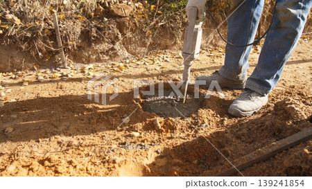 worker digs hole, power auger drills deep, worker uses auger on soil surface, compact dirt hole dug with power tool and boots, excavation performed by worker with auger for post installation worker digs hole, power auger drills deep, worker uses auger on soil surface, compact dirt hole dug with power tool and boots, excavation performed by worker with auger for post installation 139241854