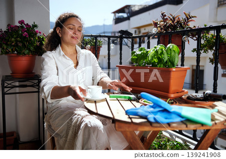 Woman Relaxing on Sunny Balcony With Potted Herbs, Coffee and Gardening Gloves 139241908