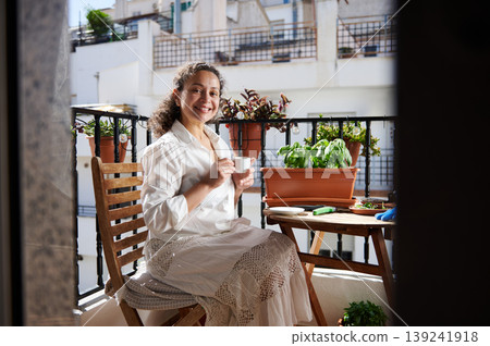 Smiling Woman Enjoying Morning Coffee on Urban Balcony With Potted Plants and Small Garden 139241918