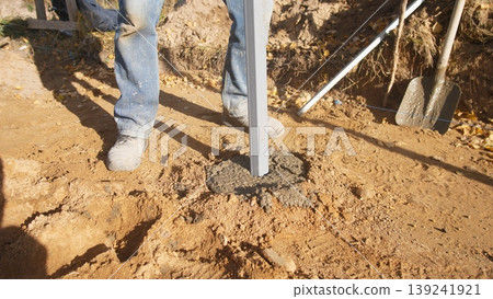worker securing post with fresh concrete at base on sunlit soil, showing poured mix, tamped surrounding earth and supportive metal post; scene conveys durable installation and finishing 139241921