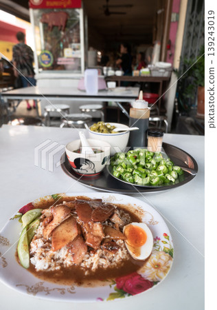 A Thai street food staple: Khao Moo Daeng. Slices of roasted red pork and crispy belly rest on jasmine rice, drenched in a rich, five-spice gravy. Accompanied by a soft-boiled egg, fresh cucumbers, 139243019