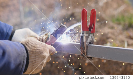 Worker Welding Clamped Joint Producing Bright Sparks And Molten Bead Closeup Shows Clamp Handles And Torch Angle Securing Two Steel Pieces For Permanent Fusion, Smoke And Light Highlight Worker Welding Clamped Joint Producing Bright Sparks And Molten Bead Closeup Shows Clamp Handles And Torch Angle Securing Two Steel Pieces For Permanent Fusion, Smoke And Light Highlight 139243150