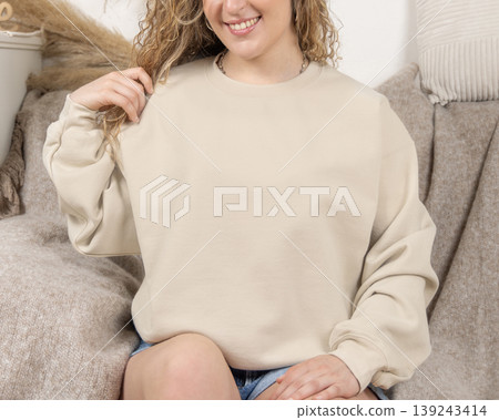 Blank beige oversized sweatshirt mockup on fair-haired young woman sitting on sofa, front view 139243414