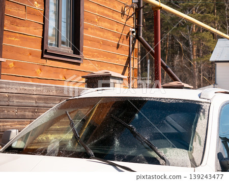Wet white SUV with raised wipers and rain droplets on windshield. Parked vehicle in front of rustic wooden log cabin against snowy rural landscape 139243477