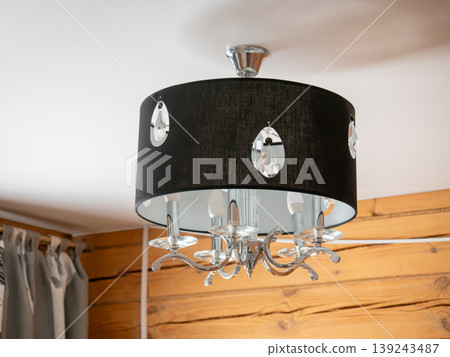 Modern crystal chandelier with black fabric shade hanging from ceiling. Stylish indoor lighting fixture for living room or bedroom. Elegant home decor and interior design element. Modern crystal chandelier with black fabric shade hanging from ceiling. Stylish indoor lighting fixture for living room or bedroom. Elegant home decor and interior design element. 139243487