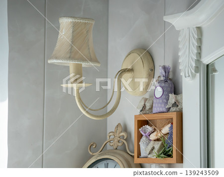 Vintage lamp mounted on wall with decorative shelf containing lavender sachets and flowers. Interior design detail with antique style elements for cozy home living and cottage decor. 139243509