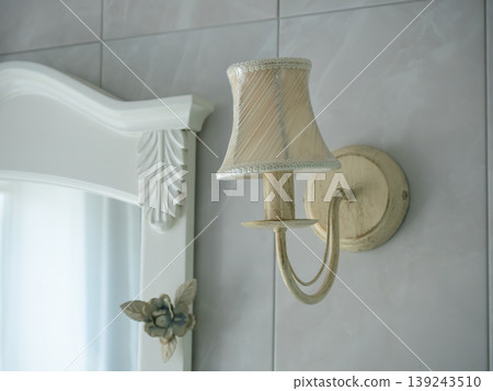 Vintage wall sconce lamp mounted on tiled bathroom wall near decorative mirror frame. Classic interior lighting fixture in elegant bedroom or restroom design. Home decoration accessory. Vintage wall sconce lamp mounted on tiled bathroom wall near decorative mirror frame. Classic interior lighting fixture in elegant bedroom or restroom design. Home decoration accessory. 139243510