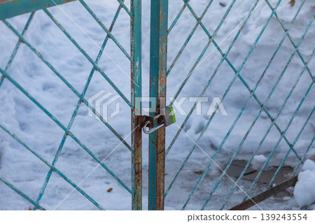 Metal gate with rusty chain link mesh locked by padlock. Secured entrance door in winter season with snow background. Concept of safety, protection, restriction, private area closure. Metal gate with rusty chain link mesh locked by padlock. Secured entrance door in winter season with snow background. Concept of safety, protection, restriction, private area closure. 139243554