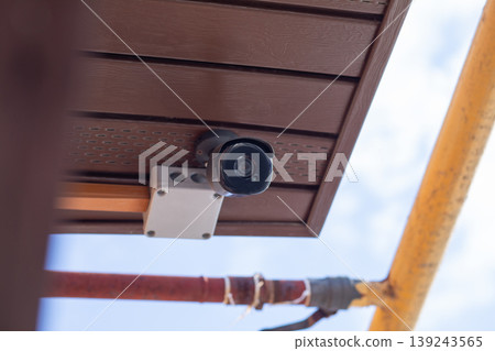 Security camera mounted on brown ceiling under roof. Surveillance system for property protection and safety monitoring. Digital video equipment for prevention of burglary and crime control. 139243565