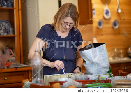 Senior woman planting seeds in pots at home. Elderly female gardener preparing seedling containers for spring season. Hobby activity, horticulture concept and sustainable home gardening. 139243688