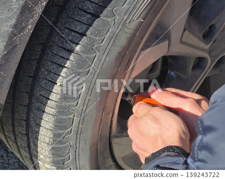 Man checking car tire pressure with orange gauge. Driver measuring air in wheel on summer day. Vehicle maintenance and safety diagnostic. Automotive repair concept for travel preparation. Man checking car tire pressure with orange gauge. Driver measuring air in wheel on summer day. Vehicle maintenance and safety diagnostic. Automotive repair concept for travel preparation. 139243722