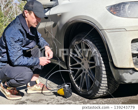 Man inflating car tire with portable electric air compressor. Technician fixing vehicle wheel pressure on roadside. Automotive maintenance, car repair and road safety service concept. 139243779
