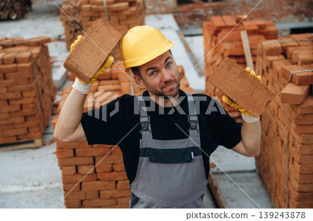 Cheerful, holding two bricks in hands. A builder is working at the construction site at daytime Cheerful, holding two bricks in hands. A builder is working at the construction site at daytime 139243878