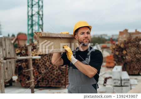 Looking to the side, moving the wooden plank. A builder is working at the construction site at daytime Looking to the side, moving the wooden plank. A builder is working at the construction site at daytime 139243907