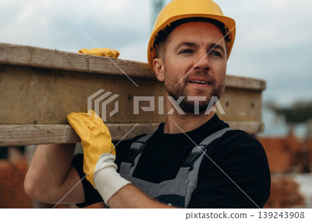 Looking to the side, moving the wooden plank. A builder is working at the construction site at daytime 139243908