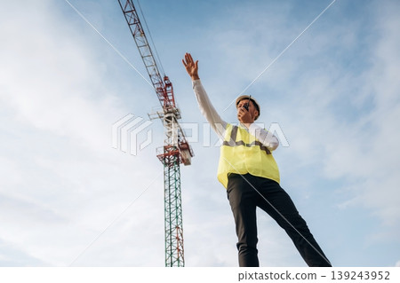 Radio communication, giving orders. Construction site worker is against building crane and sky background Radio communication, giving orders. Construction site worker is against building crane and sky background 139243952