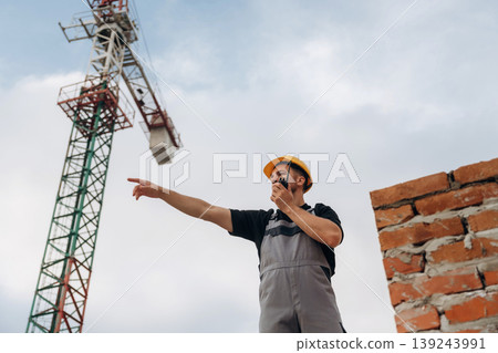Giving orders by using walkie talkie. A builder is working at the construction site at daytime Giving orders by using walkie talkie. A builder is working at the construction site at daytime 139243991