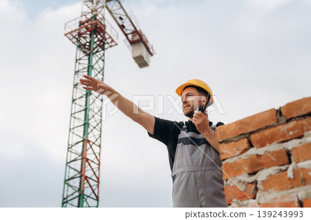 Giving orders by using walkie talkie. A builder is working at the construction site at daytime Giving orders by using walkie talkie. A builder is working at the construction site at daytime 139243993