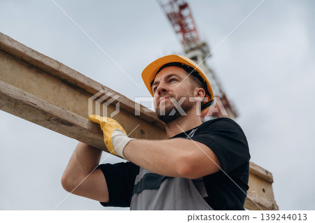 A construction worker is moving the wooden plank. In the yellow hard hat and in gloves 139244013