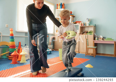 Doing balance training exercises. Preschool child with teacher during developmental activities in a playroom Doing balance training exercises. Preschool child with teacher during developmental activities in a playroom 139244293