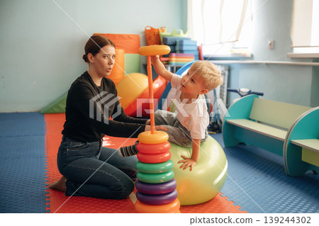 On the ball, putting rounded toys on the stick, balance. Preschool child with teacher during developmental activities in a playroom On the ball, putting rounded toys on the stick, balance. Preschool child with teacher during developmental activities in a playroom 139244302