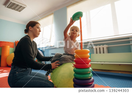 On the ball, putting rounded toys on the stick, balance. Preschool child with teacher during developmental activities in a playroom On the ball, putting rounded toys on the stick, balance. Preschool child with teacher during developmental activities in a playroom 139244303