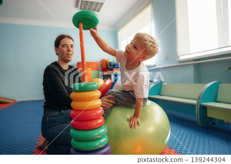 On the ball, putting rounded toys on the stick, balance. Preschool child with teacher during developmental activities in a playroom On the ball, putting rounded toys on the stick, balance. Preschool child with teacher during developmental activities in a playroom 139244304