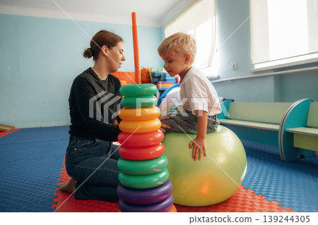 On the ball, putting rounded toys on the stick, balance. Preschool child with teacher during developmental activities in a playroom On the ball, putting rounded toys on the stick, balance. Preschool child with teacher during developmental activities in a playroom 139244305