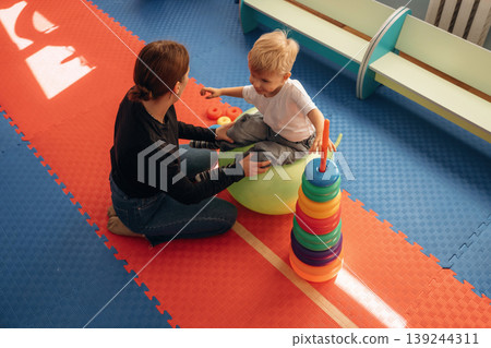 The balancing tricks on the ball. Preschool child with teacher during developmental activities in a playroom The balancing tricks on the ball. Preschool child with teacher during developmental activities in a playroom 139244311