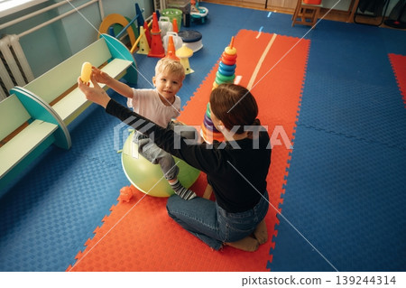 Control of the balance. Doing exercises. Preschool child with teacher during developmental activities in a playroom Control of the balance. Doing exercises. Preschool child with teacher during developmental activities in a playroom 139244314