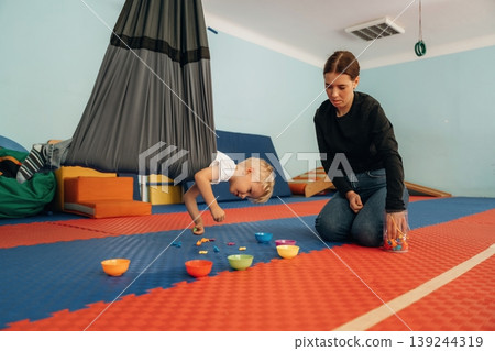 Putting the objects into bowls while hanging. Preschool child with teacher during developmental activities in a playroom Putting the objects into bowls while hanging. Preschool child with teacher during developmental activities in a playroom 139244319