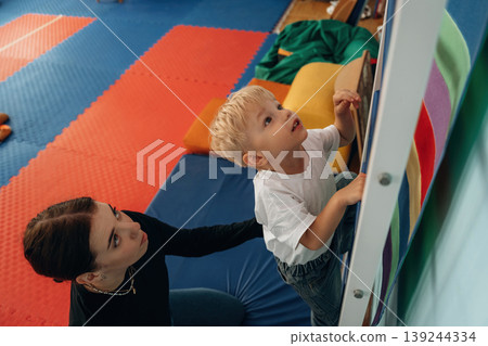 Climbing the ladder that is on the wall. Preschool child with teacher during developmental activities in a playroom Climbing the ladder that is on the wall. Preschool child with teacher during developmental activities in a playroom 139244334